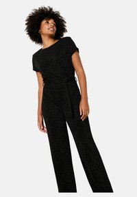 Melna kombinezona uzvalks ar īsām piedurknēm, apaļu kakla izgriezumu un jostu vidukļa daļā. Audumam ir smalks spīduma efekts, radot struktūru.