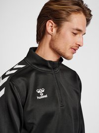 Sort halv-zip sportsjakke med hvide detaljer. Har en høj krave, glat tekstur og et logo på brystet. Kontraststriber på skuldrene.