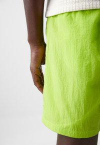 Lime green shorts in a lightweight, wrinkled fabric with a straight cut and stitched hem. Visible hand holding the shorts at the side.