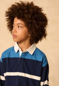 Tamsiai mėlynas polo marškinėlis su mėlynomis juostelėmis ir baltais akcentais, su apykakle ir laisvo kirpimo, dėvimas vaikų su garbanotais plaukais.