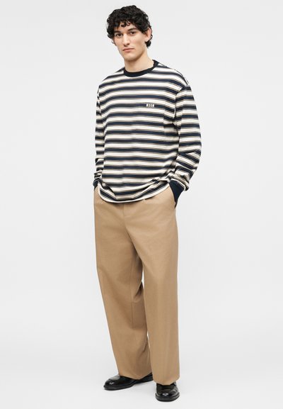 Νεαρός άνδρας στέκεται με τα χέρια στις τσέπες, φοράει μπλε ναυτικό και λευκό ριγέ πουκάμισο με μακριά μανίκια, μπεζ παντελόνι με φαρδιά πόδια και μαύρα παπούτσια.