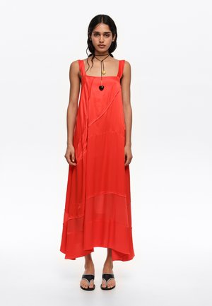 Mujer con vestido rojo sin mangas, fluido, con escote cuadrado y detalles en capas, sandalias negras y collar con colgante de corazón negro y dorado.