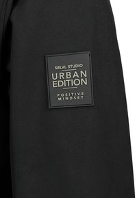 Zwarte stof met een rechthoekig label. De tekst luidt "SBLVL STUDIO URBAN EDITION" en "POSITIVE MINDSET." Glad oppervlak, modern ontwerp.