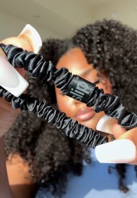 Svarta scrunchies av glatt satinmaterial, med en samlad textur och en liten logodetalj på en av dem. Två hålls mot en suddig bakgrund.