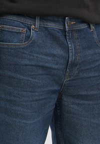 Temno modri denim jeans z gladko teksturo, okrašeni s bakrenimi zakovicami, šivi in standardnimi žepi. Brez vidnih logotipov.