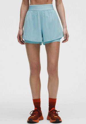 Kvinde iført lyseblå lagdelte atletikshorts, orange sokker og matchende orange løbesko, stående mod en ensfarvet baggrund.