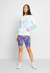 Šarena tie-dye dukserica u pastelnim nijansama s prednjim džepom, uparena s prilagođenim biciklističkim šortsevima s apstraktnim uzorkom u ljubičastoj i ružičastoj boji.