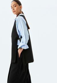 Chaleco negro a medida con hebilla de plata ajustable, llevado sobre una camisa de cuello azul claro. El chaleco tiene un corte relajado y bolsillos laterales.