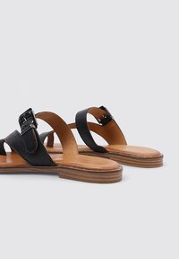 Sandalias de cuero negro con una suela plana en tono bronceado. Presentan tiras cruzadas con un acento de hebilla, textura suave y diseño minimalista.
