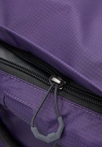 Sac à dos en tissu violet avec un extérieur texturé, fermeture éclair noire et un tirant sécurisé. Les coutures visibles et le compartiment avec logo mettent en avant la durabilité.