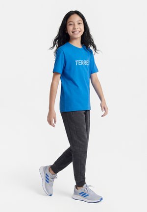 Junge Person mit langen dunklen Haaren lächelt, trägt ein blaues TERREX T-Shirt, dunkelgraue Jogginghosen und weiße Sneakers mit blauen Streifen, geht.