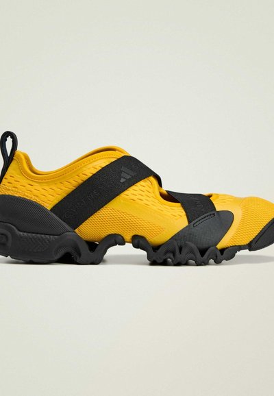 Zapatillas deportivas amarillas con parte superior texturizada, correa negra y suela ondulada de goma. Incluyen una lengüeta para tirar y malla para la transpirabilidad.