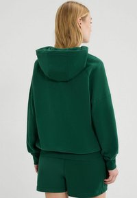 Grüne Kapuzenjacke mit lockerer Passform, die eine Kapuze, weite Ärmel und gerippte Bündchen aufweist. Der Stoff ist glatt und sorgt für eine weiche Textur.