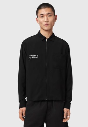 Jonge man die een zwart shirt met ritssluiting draagt, met kraag, borstzak en witte geborduurde tekst op de linkerborst.