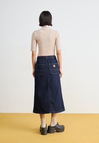 Denim maksi suknja u tamnoplavoj boji s kontrastnim šavovima, dva stražnja džepa i oznakom; u kombinaciji s bež majicom i crnim klomama.