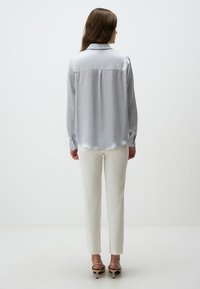Hellgraue Satinbluse mit einem Rückeneinsatz, Button-Down-Kragen und langen Ärmeln, kombiniert mit hellen Hosen und Riemchensandalen.
