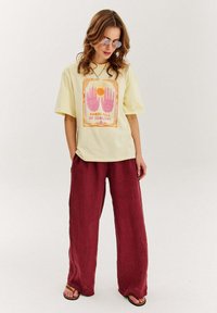 Žluté bavlněné tričko s grafickým potiskem rukou a slunce, spárované s volnými burgundskými lněnými kalhotami a černými sandály.