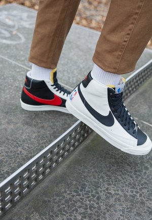 Person iført mismatchede Nike high-top sneakers, den ene sort og hvid med NBA-logo, den anden sort med rød swoosh, træder på et metal bordtennisbord.