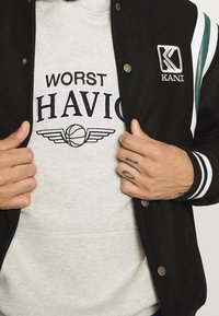 Zwarte varsityjack met groene en witte streepaccenten, gelaagd over een lichtgrijze hoodie met de tekst "WORST HAVIOC" en een basketbalgrafiek.