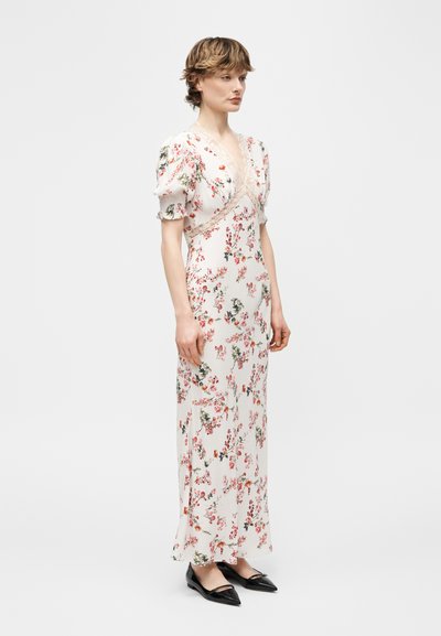 Γυναίκα που φοράει λευκό μακρύ φόρεμα με λουλουδάτο μοτίβο, φουσκωτά μανίκια και δαντελένιο τελείωμα, σε συνδυασμό με μαύρα μυτερά επίπεδα παπούτσια, στέκεται μπροστά σε ένα απλό φόντο.