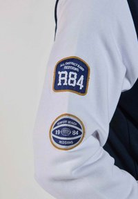 Chemise blanche à manches longues avec des accents bleu marine. Deux écussons brodés : l'un avec "R84" et l'autre avec "Newport Beach 1934" dans un design circulaire.