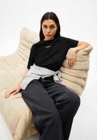 Schwarzes Kurzarmhemd mit einem weißen Logo, kombiniert mit dunkelgrauen, weiten Jeans und einem hellgrauen Pullover, der um die Taille gebunden ist, auf einem beigen Sofa.