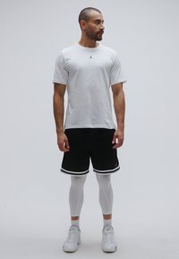 Λευκό πουκάμισο με κοντά μανίκια και μαύρο λογότυπο, μαύρο σόρτς με λευκή περιγραφή, λευκά κολάν και λευκά αθλητικά παπούτσια. Το μοντέλο στέκεται μπροστά σε ουδέτερο φόντο.