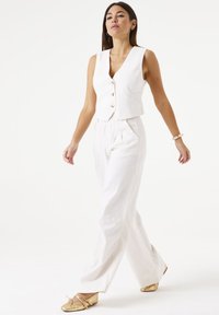 Witte linnen mouwloze vest met ronde knopen, gecombineerd met hoge witte linnen broeken. Model draagt gouden platte schoenen, in looppose.