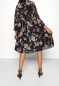 Black floral dress with 3/4 sleeves, featuring white and purple flower patterns, flowing silhouette, and knee-length hem. Worn with black heels.