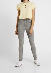 Camiseta amarilla recortada con mangas enrolladas, combinada con vaqueros ajustados grises de cintura alta y zapatillas blancas con detalles en negro y rojo.