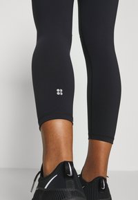 Schwarze Sportleggings mit einer eng anliegenden Passform und einer glatten Textur, versehen mit einem kleinen silbernen Logo über dem Knöchel. Schwarze Sportschuhe.