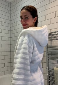 Peignoir de bain à capuche blanc avec des rayures texturées, fabriqué en matériau doux, doté d'une taille à nouer et d'un fond de murs de salle de bains carrelés.