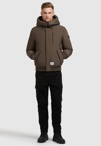 Bruine gewatteerde jas met een hoge kraag, trekkoordcapuchon, ritszakken en zwarte cargobroek. Draag met zwarte laarzen.