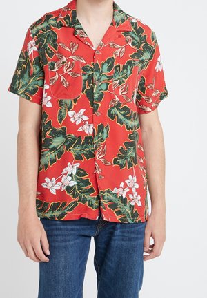 Homme portant une chemise rouge à manches courtes avec de grandes feuilles vertes et des motifs de fleurs blanches, associée à un jean bleu.