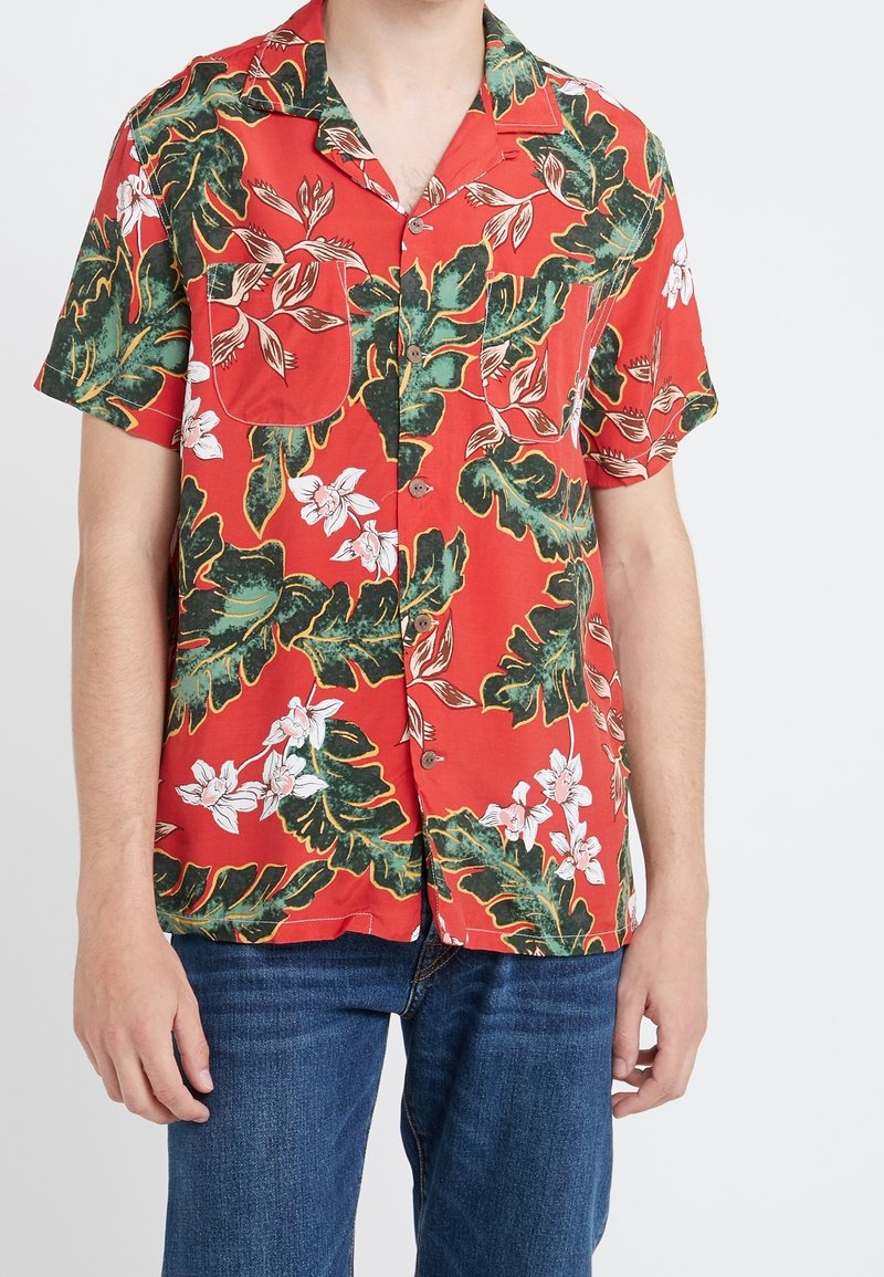 Homme portant une chemise rouge à manches courtes avec de grandes feuilles vertes et des motifs de fleurs blanches, associée à un jean bleu.
