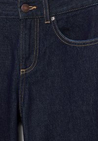 Dettaglio dei jeans in denim blu scuro che mostra la tasca anteriore, il rivetto in rame, il bottone con incisione del marchio e le cuciture arancioni a contrasto.
