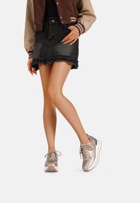 Sneaker in un leggero argento metallizzato con una texture liscia, forma arrotondata e design con lacci. Indossati con una gonna di jeans nera con orlo sfrangiato.