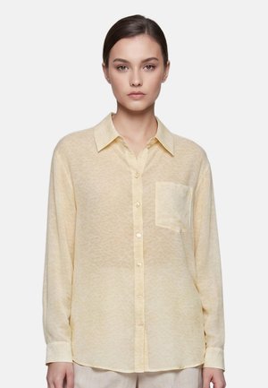 Giovane donna con capelli scuri che indossa una camicia giallo chiaro con bottoni e una tasca sul petto, in piedi davanti a uno sfondo bianco uniforme.
