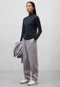 Ciemnoszary sweter z golfem w zestawie z jasnoszarymi, plisowanymi spodniami o szerokich nogawkach, trzymając dopasowaną kurtkę i nosząc białe sneakersy.