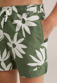 Zaļie peldšorti ar lielu baltu ziedu rakstu, elastīgu jostu, auklu un sānu kabatām. Iespējots mazs logo uz apakšmalas.