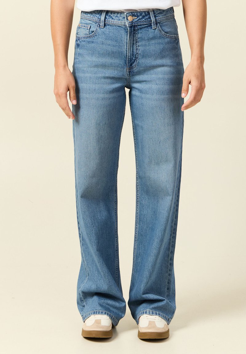 Person, die hellblaue Jeans mit hohem Bund und geradem Bein sowie weiße Turnschuhe trägt, steht vor einem schlichten, hellen Hintergrund.