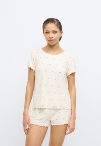 Ελαφρύ σετ πυτζάμες από κρεμ βαμβάκι με κοντά μανίκια και ριμπ ύφασμα· διαθέτει μικρά ροζ καρδιές και κυματιστό περίγραμμα στις άκρες.