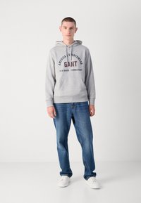 Sudadera gris con el logo y texto "GANT", bolsillo tipo canguro, puños de canalé, combinada con jeans azules y zapatillas blancas sobre un fondo neutro.