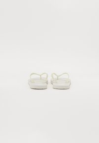 Hvite gummisandaler med tynne, bølgende stropper og en flat såle. Designet har en minimalistisk form og en jevn tekstur.