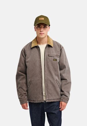 Jeune homme portant une veste marron avec un col beige sur une chemise claire, un jean foncé et une casquette de baseball vert olive, debout devant un fond uni.