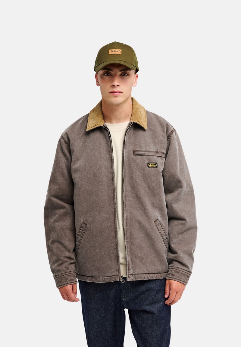 Jeune homme portant une veste marron avec un col beige sur une chemise claire, un jean foncé et une casquette de baseball vert olive, debout devant un fond uni.