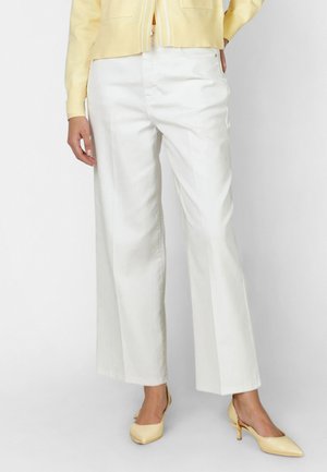 Frau trägt weiße, weit geschnittene Hosen, gelben Cardigan und gelbe Schuhe mit niedrigem Absatz, steht vor einem schlichten Hintergrund.