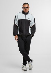 Vyras, vilkintis juodą ir šviesiai pilką Sergio Tacchini striukę, juodas kelnes, baltus sportbačius ir akinius nuo saulės, stovi sukėlęs kojas.