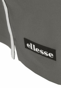 Grijze stof met een gladde textuur, voorzien van een zwart label met witte letters waarop "ellesse" staat, en een contrasterende witte bies.