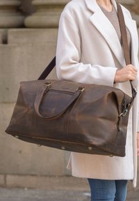 Skórzana torba podróżna w ciemnym brązie, z podwójnymi uchwytami, regulowanym paskiem na ramię, boczną kieszenią i mosiężnymi akcentami. Gładka faktura, duży rozmiar.